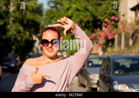 Sourire de jeune fille faisant photo frame avec ses mains. Elle est debout dans la rue, avec des voitures en arrière-plan. Chaude journée ensoleillée. Banque D'Images