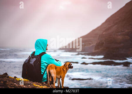 Homme avec un chien en face de paysage littoral rural avec des montagnes et des vagues venant et rayons de soleil à travers les nuages. L'île de Santo Antao, Cap Vert Banque D'Images