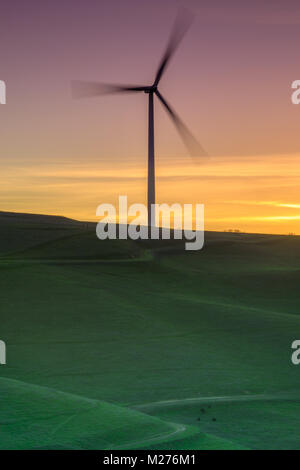 Lone wind turbine tournant sur une colline herbeuse au coucher du soleil. Banque D'Images