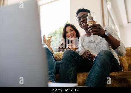 L'Interracial couple sitting on sofa avec verres de vin et de manger du popcorn pendant la prise de film sur l'ordinateur portable dans la salle de séjour. Banque D'Images