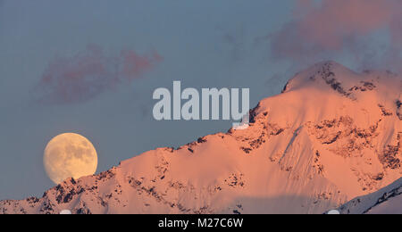 Une presque pleine lune se lève derrière pic de l'Aigle baigné dans alpine glow minutes avant le coucher du soleil à la mi-printemps dans les montagnes de Chugach, Southcentral Alaska. Banque D'Images