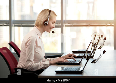 Adorable smiling female callcenter agent. Femme blonde mature vue du côté des travailleurs du service à la clientèle, centre d'appel opérateur souriant avec casque téléphonique. Banque D'Images