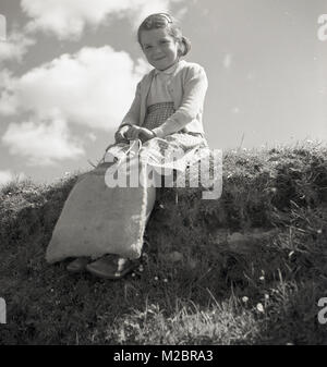 Années 1950, historique, un portrait par Allan J'encaisse de une douce jeune fille de la campagne irlandaise assis sur une banque d'herbe portant son sac de jute. Banque D'Images