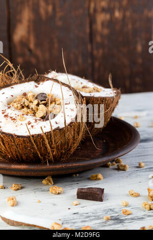 Petit-déjeuner sain dans la noix de coco noeud sur fond blanc. Le yogourt dans un bol de noix de coco avec des flocons de noix de coco, chocolat et muesli. Vue de dessus, des laïcs, des frais généraux Banque D'Images