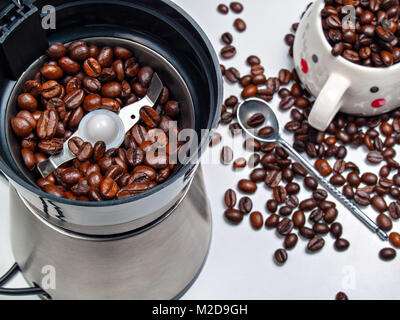 Moulin à café électrique avec grains de café torréfié à l'intérieur et les grains de café éparpillés sur une table avec une cuillère en métal à partir de ci-dessus et une tasse remplie de cof Banque D'Images