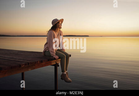 Caucasian woman sitting on dock du lac en admirant le coucher du soleil Banque D'Images