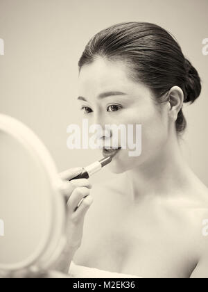 Belle jeune femme asiatique à la recherche dans le miroir tout en appliquant le rouge à lèvres sur des lèvres, noir et blanc, style rétro. Banque D'Images