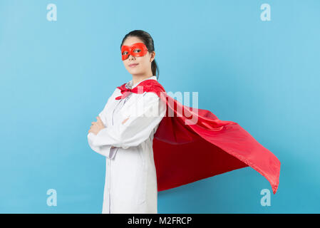 Confiance professionnelle femme médecin wearing superhero costume avec masque debout dans le fond bleu et les mains de mettre sur la poitrine face à la caméra. Banque D'Images