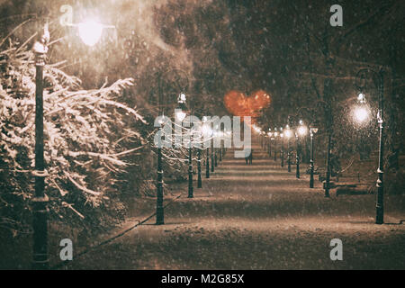 Couple in love sur une date romantique promenades à travers la nuit de neige winter park alley avec de belles lanternes couverte de neige au cœur formé par les branches d'arbres sur la St Valentin day Banque D'Images