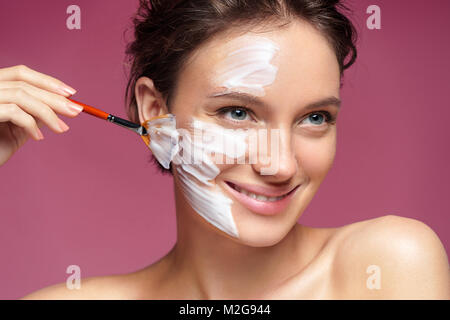 Femme avec masque facial hydratant et les mains de l'esthéticienne sur fond rose. Photo de jolie femme reçoit les traitements de spa. Beauté et peau c Banque D'Images