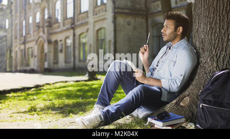 Guy multinationales assis sous arbre dans park écrit idée créative dans sketchbook Banque D'Images