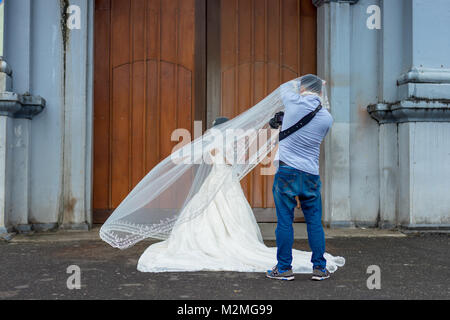 Photographe de mariage habillé décontracté prend des photos d'une épouse dans une magnifique robe blanche aux portes de son église. Banque D'Images