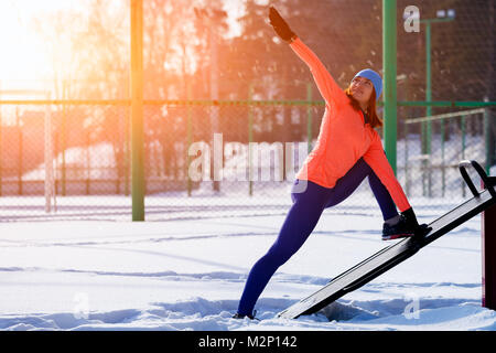 Jeune femme dans le bleu de l'hat, chandail orange et de l'élan avant l'exécution s'étend sur un terrain de sport sur un jour d'hiver lumineux Banque D'Images