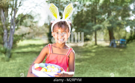 Mignon et belle African American girl wearing bunny ears, regarde en souriant de l'appareil photo tout en maintenant l'Easter basket plein d'œufs. Fille est assise dans la surenchère du parc Banque D'Images