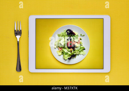 Une photo, prise par moi-même, d'une salade verte dans une assiette blanche sur fond jaune dans l'écran d'une tablette, sur un fond jaune à côté d'un f Banque D'Images