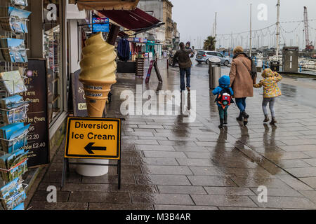 Femme avec deux enfants à marcher le long du front de mer de Torquay par temps humide au début de la petites vacances de février 2018. Rack de mer y compris l'attirail de cartes postales et de la crème glacée sont sur l'affichage en dépit du mauvais temps. Torbay Torquay, Devon, UK., Février 2018. Banque D'Images