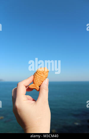 Main tenant une mini gaufre en forme de poisson avec de la crème anglaise le remplissage avec la mer en arrière-plan journée ensoleillée, Corée du Sud Banque D'Images