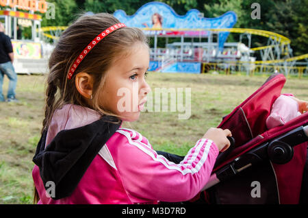 Petite fille en rose veston sport avec serre-tête tenant un landau dans l'arrière-plan attractions Banque D'Images