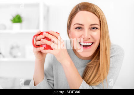 Une jolie jeune femme tenant une tasse rouge assis à une table dans un bureau blanc. Pause café. Banque D'Images