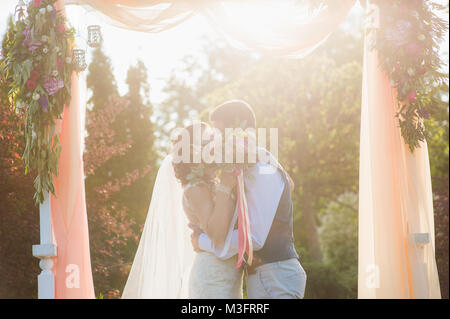 Marié à la femme et l'homme s'embrassent en été vert nature contexte au cours de la cérémonie du mariage. Heureux couple marié à lumière douce de soleil couchant. Banque D'Images