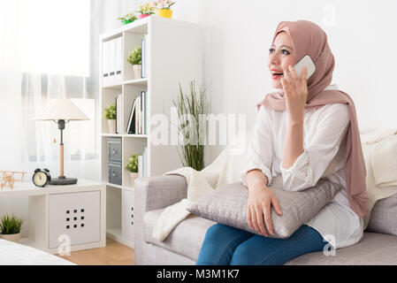 Happy happy pretty muslim woman sitting on sofa et relaxant à l'aide de mobile cell phone appelant à un ami pendant les vacances. vacances chat Banque D'Images