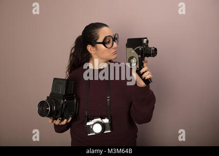 La photographie féminine nerdy buff portant de grandes lunettes avec trois caméras vintage brandissant une caméra vidéo et regarder il with copy space Banque D'Images