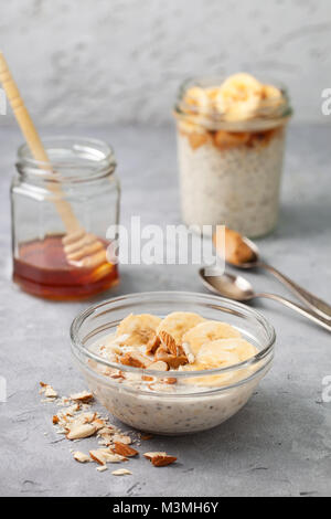 Régime alimentaire sain petit déjeuner. nuit avoine avec chia seeds, bananes, beurre d'arachide, de miel dans un bocal en verre sur un fond de béton gris Banque D'Images