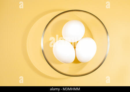 Trois œufs blancs sont couché dans une tasse en verre transparent ronde sur un fond jaune. Vue d'en haut. Banque D'Images