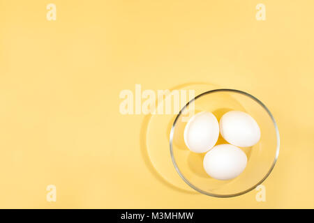 Trois œufs blancs sont couché dans une tasse en verre transparent ronde en bas à droite l'un fond jaune et copiez l'espace. Vue d'en haut. Banque D'Images