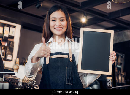 Asian female barista de porter un tablier jean Thumbs up at blank blackboard menu café au comptoir bar avec smile face,cafe service concept,propriétaire de l'entreprise star Banque D'Images