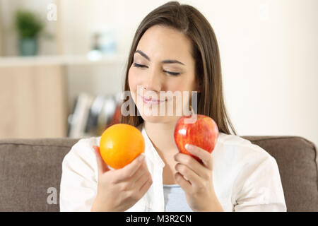 Vue avant, portrait d'une femme de décider entre un douteux et apple orange assis sur un canapé à la maison Banque D'Images