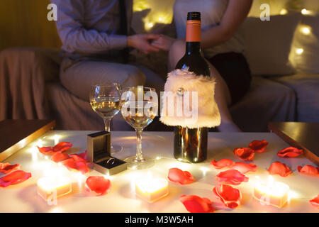 ​Couple in love sur canapé célébrant l'engagement à la maison le soir. L'accent sur la table avec des verres de vin et de cadeaux. Banque D'Images