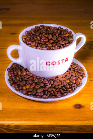 Costa Coffee : logo sur un très grand verre (avec doubles poignées), assis sur une soucoupe, pleine de grains de café rôti entier, sur un ancien plan de pin. Angleterre, Royaume-Uni. Banque D'Images