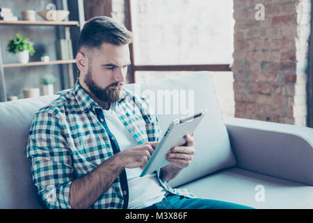 À l'aide d'homme barbu, travaillant sur PC tablette numérique sans fil, assis sur le canapé dans le salon, avec l'expression grave Banque D'Images