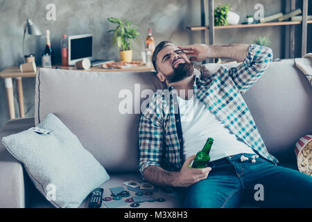 Beau et élégant, l'homme souffrant de maux de tête après la nuit de la partie le matin, tenant une bouteille de bière à la main, sitting on sofa in living room Banque D'Images