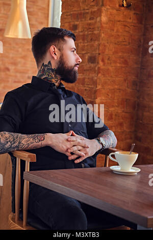 Un homme avec des bras tatoués se reposant à la table avec une tasse de café Banque D'Images