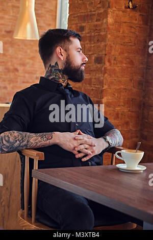 Un homme avec des bras tatoués se reposant à la table avec une tasse de café Banque D'Images