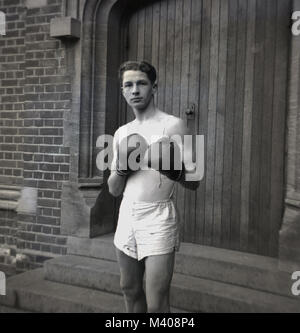 1950, historiques...le "noble art"...un jeune élève de l'école d'une grande école en bois porte à Bancrofts, Woodford Green, Angleterre, Royaume-Uni, portant une veste de sport et des courts-circuits et des gants de boxe et de prendre une pose ou de frappe monétaire. La boxe était un sport enseigné tant en état et de public (écoles privées) en ce moment, et en dehors de la santé et forme physique, les élèves ont appris que les combats ont eu lieu dans un ring, pas sur une rue. Banque D'Images