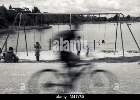 Une photo en noir et blanc d'un cycliste passant rapidement devant les enfants jouant sur les balançoires dans un parc Banque D'Images