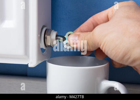 Close-up of Person's Hand de tourner la vanne de purge du radiateur pour libérer l'air avec tasse à la maison Banque D'Images