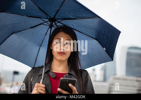 Businesswoman in city holding umbrella looking at smartphone Banque D'Images