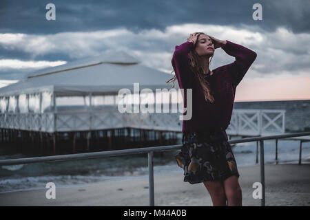 Jeune femme avec les mains dans les cheveux longs appuyé contre beach pier au crépuscule, Odessa, Ukraine Banque D'Images