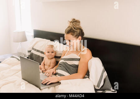 Portrait of cute baby girl sitting jusqu'au lit alors que mère using laptop Banque D'Images