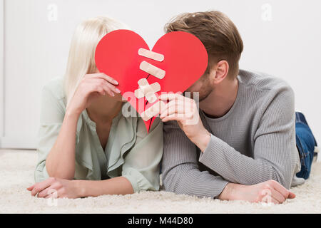 Couple Lying On Carpet Holding Paper Cœur rouge fixe avec bandage en plâtre Banque D'Images