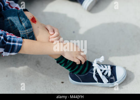 Enfant blessé assis sur le sol en béton et en regardant son genou écorché Banque D'Images