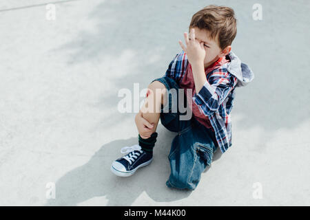 Petit garçon blessé assis sur un sol en béton avec un genou écorché Banque D'Images