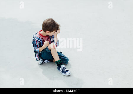Petit garçon blessé assis sur un sol en béton avec un genou saignement Banque D'Images