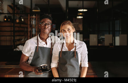 Heureux propriétaire d'une boutique de café couple standing à l'intérieur de leur boutique. L'homme et la femme des baristas se tenant debout à l'intérieur leur café portant un tablier avec l'homme qui tient le café Banque D'Images