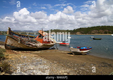 Vieux bateau de pêche chalutier tourné sur un côté, ruiné et pourrir sur un rivage rocheux par la banque de la rivière Mira. De plus petits bateaux autour. Ciel nuageux bleu vif. Banque D'Images