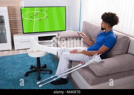 Jeune homme à la jambe cassée Watching Football Match à la télévision Banque D'Images